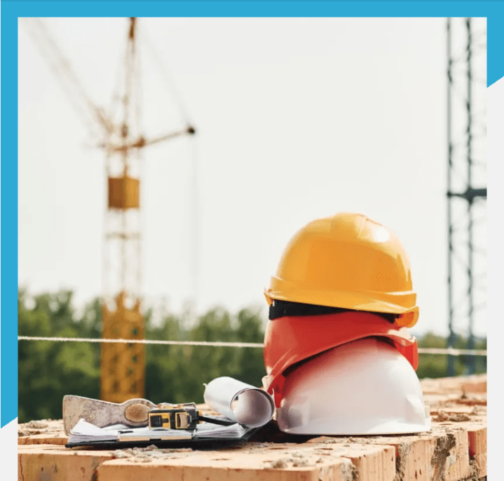 Stack of construction safety helmets on a brick wall with building plans and measuring tape, construction crane and scaffolding in the background. Professional roofing and construction services in progress.