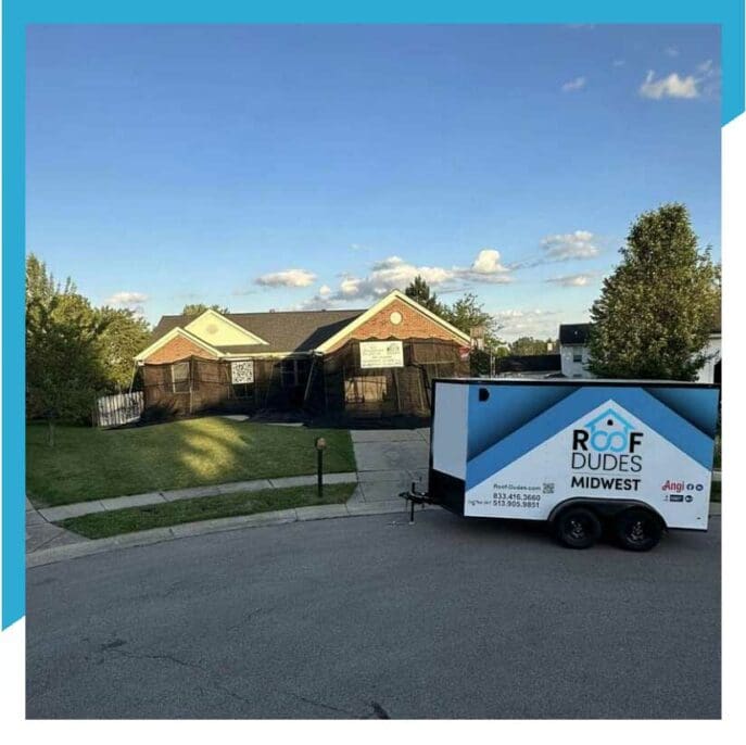 Roof Dudes Midwest trailer parked in front of a residential home undergoing roof replacement with black safety netting, clear blue sky background. Professional residential roofing services in the Midwest region.