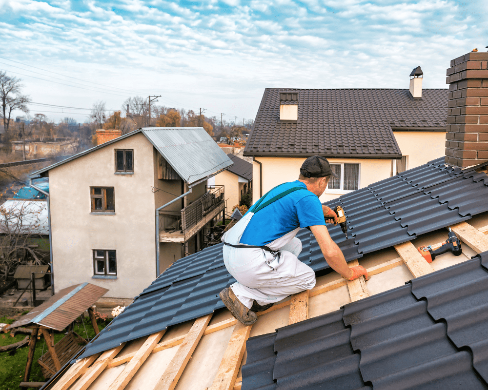 A roofer in a blue shirt and white overalls installs black metal roofing panels on a sloped roof using a power drill. The wooden framework beneath the panels is visible, showing the process of securing the new roof.