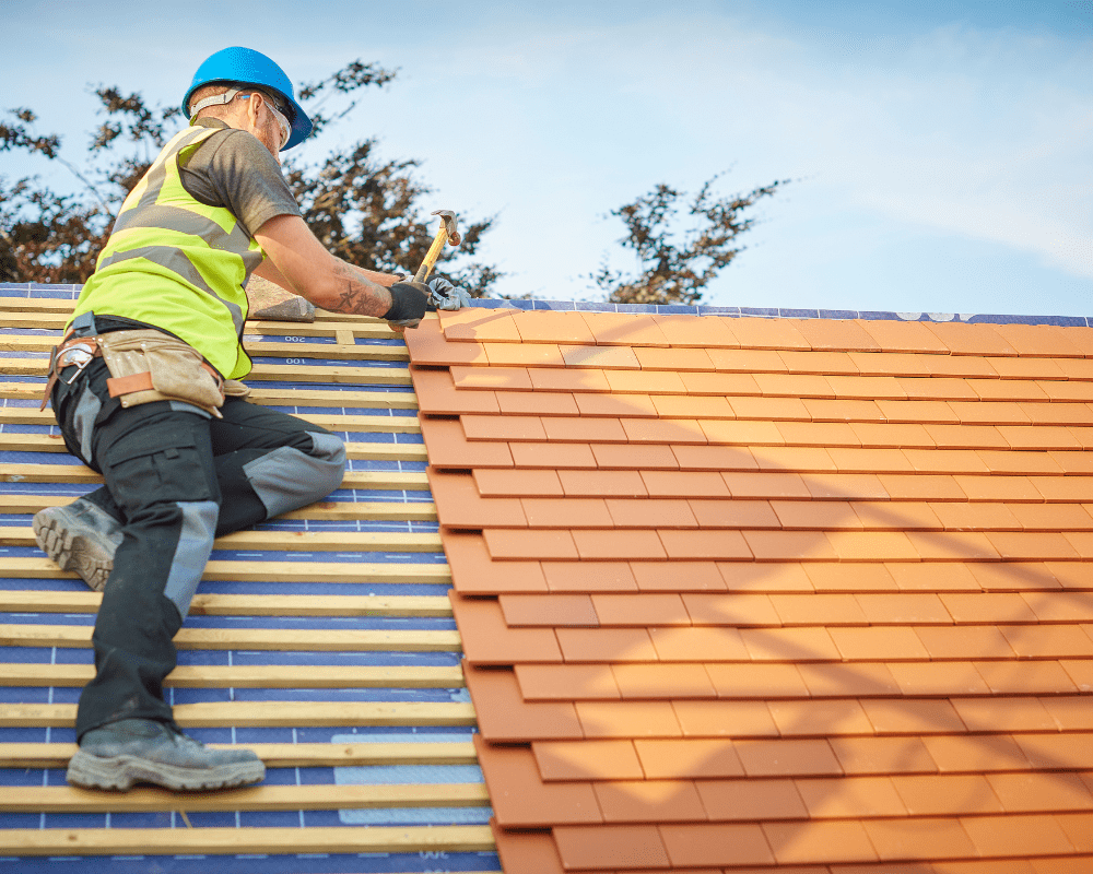 A residential roofing contractor wearing a blue hard hat, high-visibility vest, and tool belt installs terracotta-colored roof tiles on a sloped roof. He uses a hammer to secure the tiles to the wooden framework beneath. The background features a clear blue sky and tree branches.